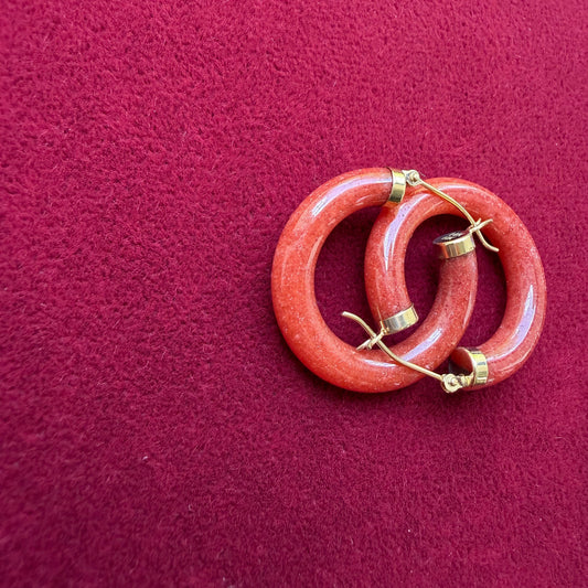 A pair of red jade hoop earrings with 14k gold accents, displayed on a textured dark red velvet surface.