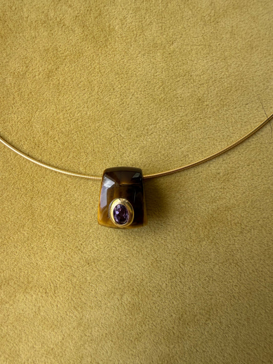 A tiger's eye pendant with a faceted amethyst stone in a gold bezel setting, suspended on a thin gold necklace against a textured olive green background.