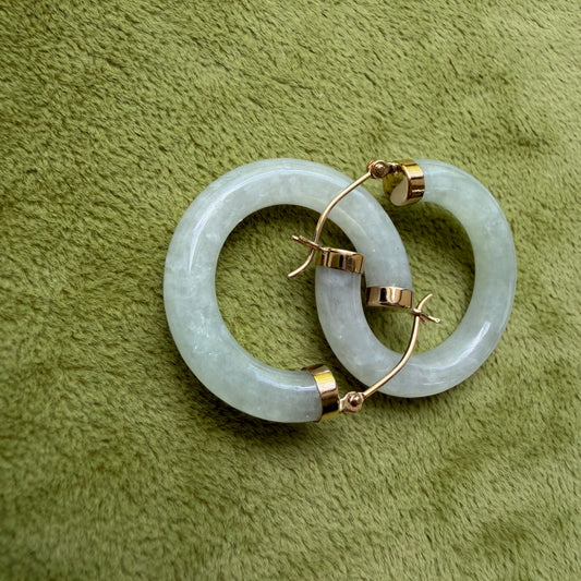 A pair of light green jade hoops with 14k gold hooks, displayed on a textured olive green fabric background.