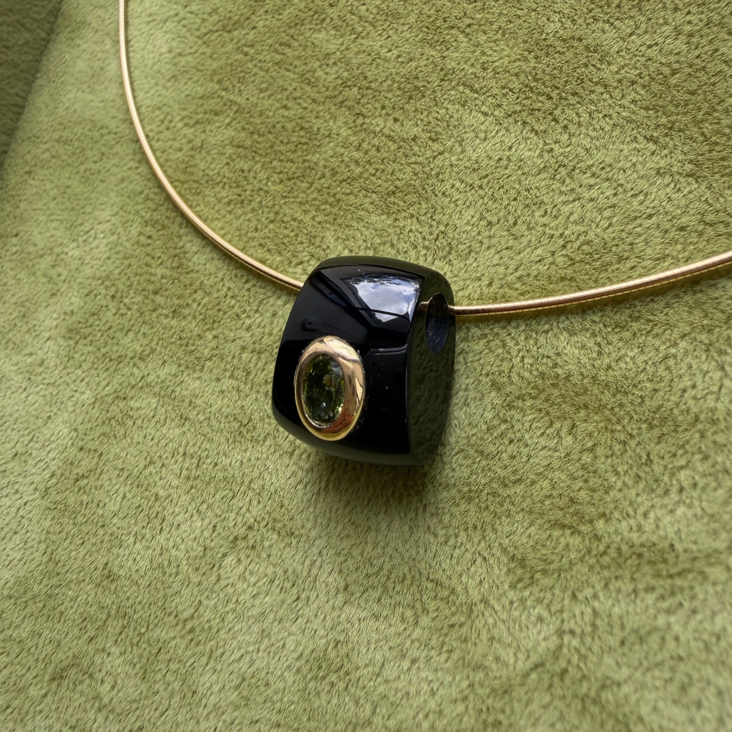 A close-up of a black onyx pendant with a 14k gold bezel featuring a peridot stone, threaded onto a thin gold necklace against a textured olive green background.