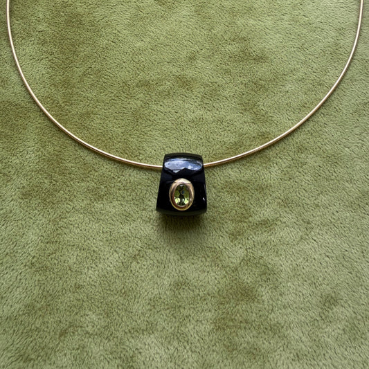 A black onyx pendant featuring a central peridot, displayed on a textured olive green background.