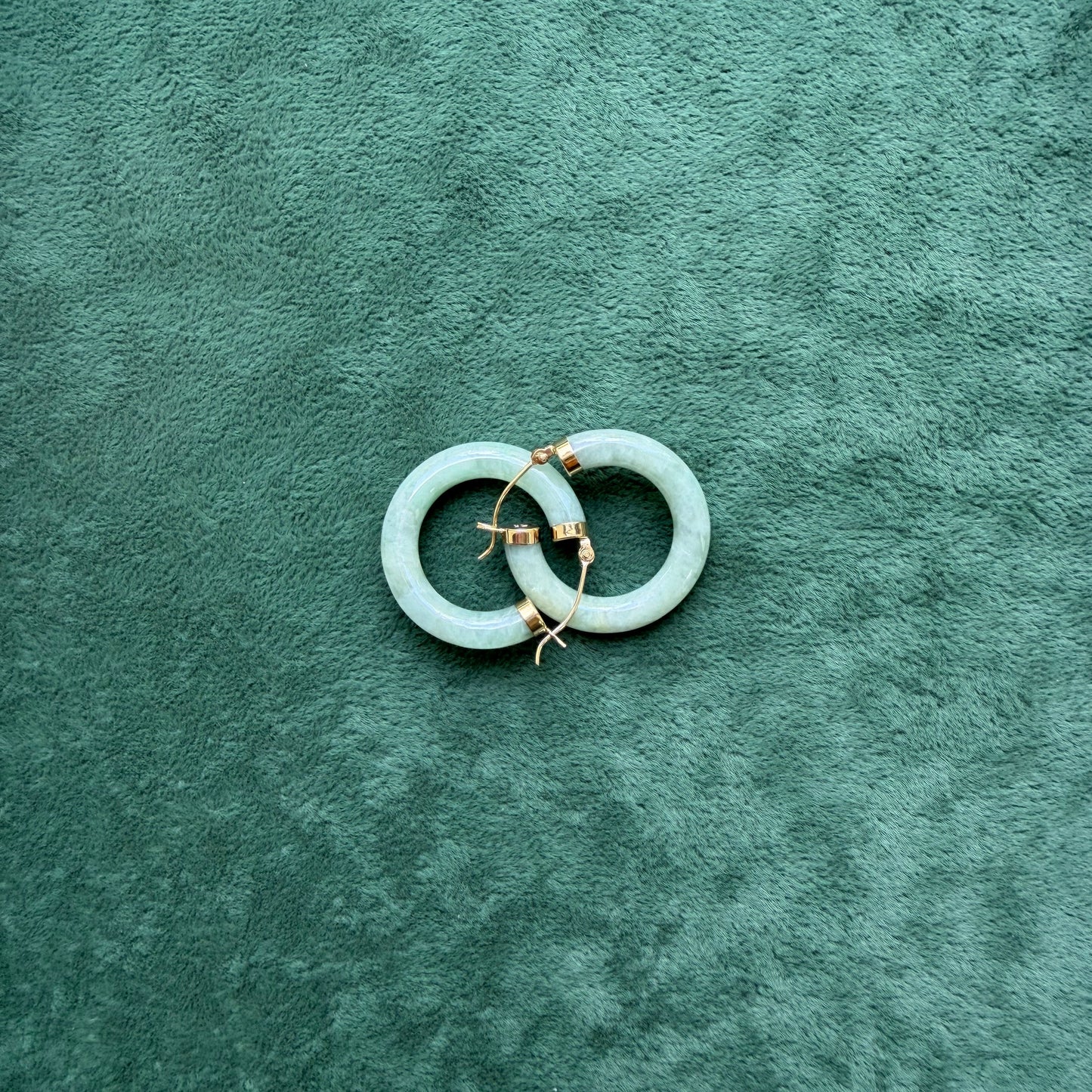 Two green jade hoops with 14k gold hooks and end caps, displayed on a textured dark green velvet surface.