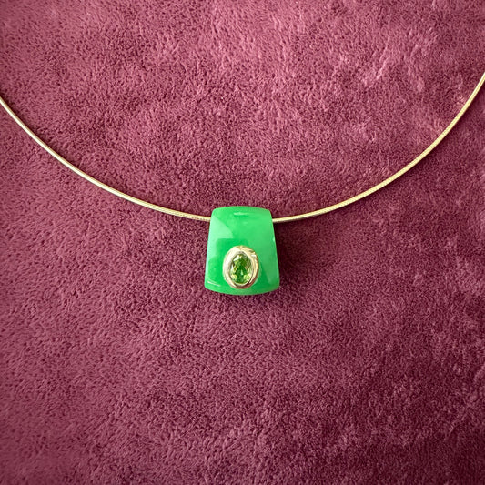 A green jade pendant with a 14k solid gold bezel hugging a peridot stone, threaded onto a thin gold wire, set against a textured dark purple background