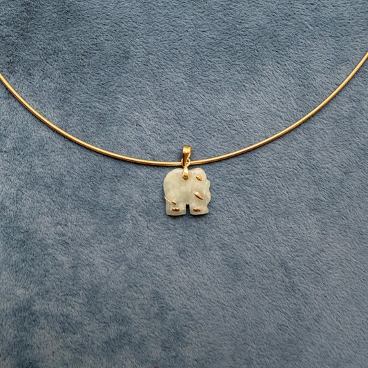 A delicate gold wire necklace with a small, light green jade carved elephant pendant, laid flat on a textured blue-gray fabric background.