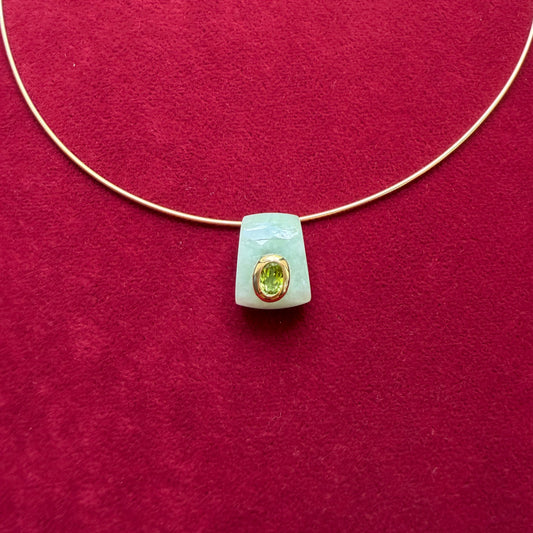 A delicate gold wire necklace with a pale green jade pendant featuring a gold bezel-set peridot, displayed on a textured dark red background.