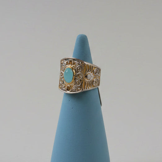 An ornate vintage-style ring with an aquamarine cabochon and filigree detailing, displayed on a blue cone stand against a neutral gray background.