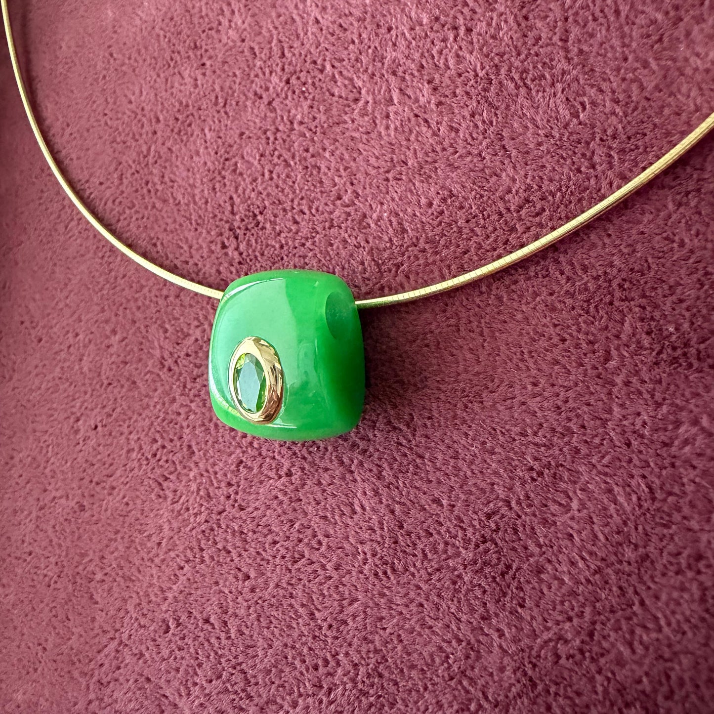 A side view of a green jade slider pendant with a 14k gold-toned bezel hugging a peridot stone at the center, threaded on a gold cord against a textured dark purple background.