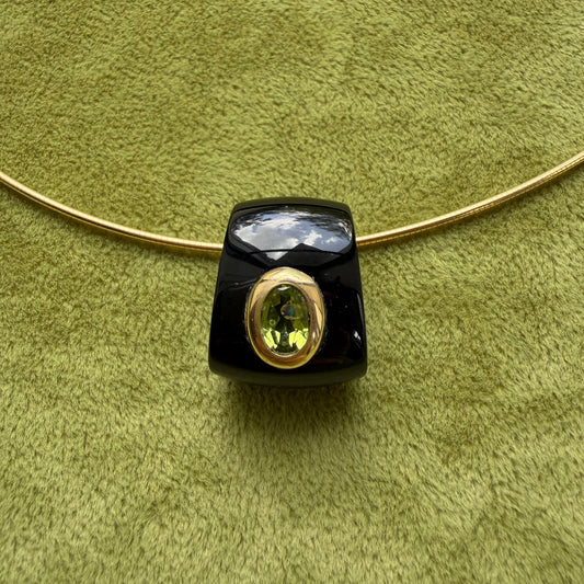 A black onyx pendant with a 14k gold bezel-set peridot, shown on a textured olive green background with a gold chain.
