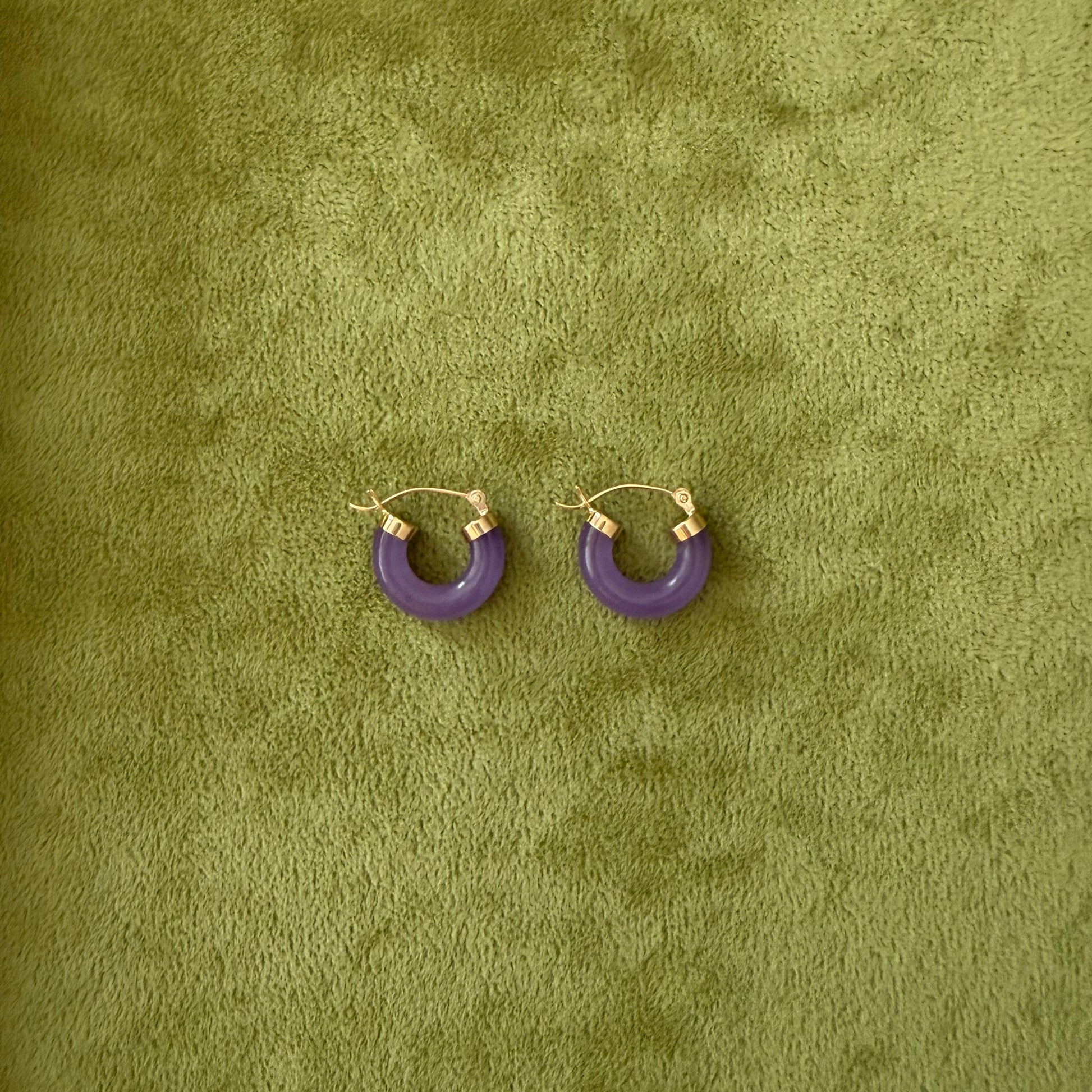 A pair of small lavender jade hoop earrings with 14k gold hooks, displayed on a textured olive green surface.