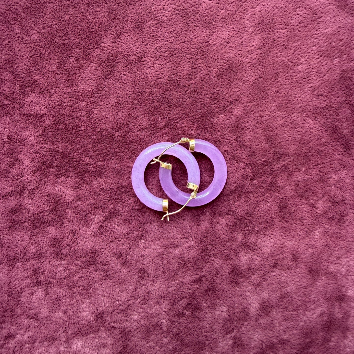 Two lavender jade hoops with 14k gold hooks and end caps, displayed on a textured, deep purple velvet surface.
