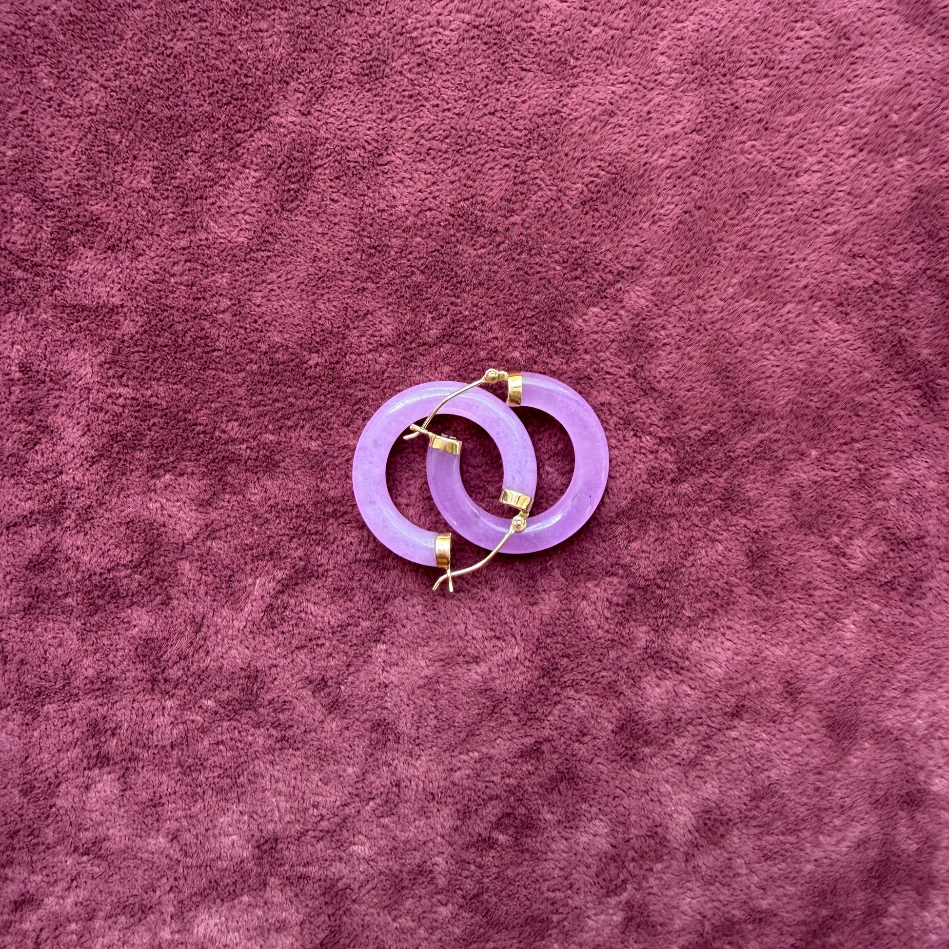 Two lavender jade hoops with 14k gold hooks and end caps, displayed on a textured, deep purple velvet surface.