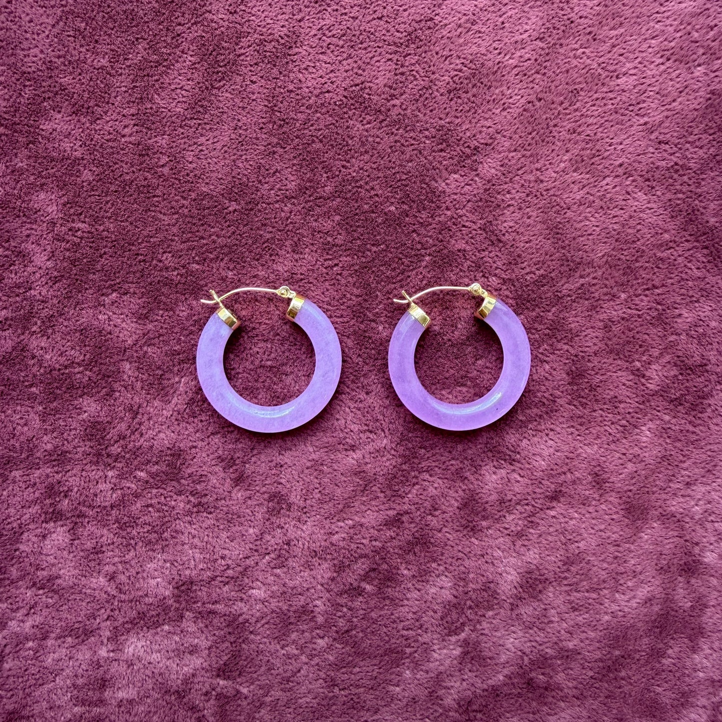 A pair of lavender jade hoop earrings with 14k gold fishhook backings, displayed on a textured purple velvet surface.