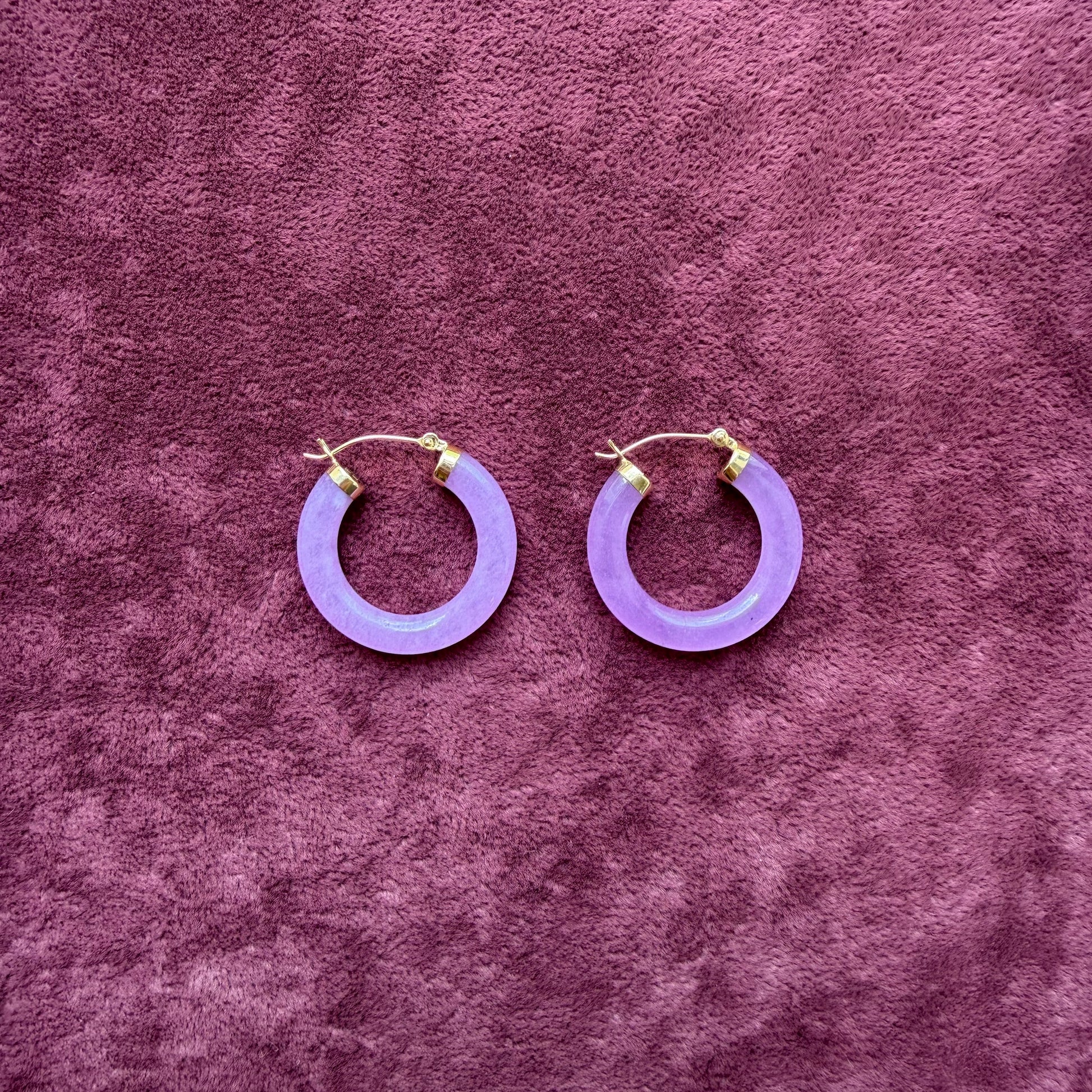 A pair of lavender jade hoop earrings with 14k gold fishhook backings, displayed on a textured purple velvet surface.