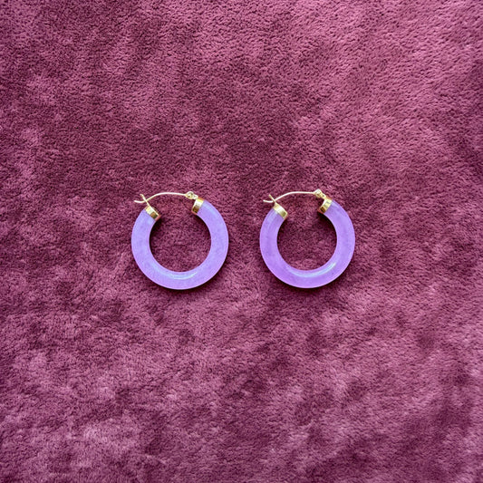 A pair of lavender jade hoop earrings with 14k gold fishhook backings, displayed on a textured purple velvet surface.