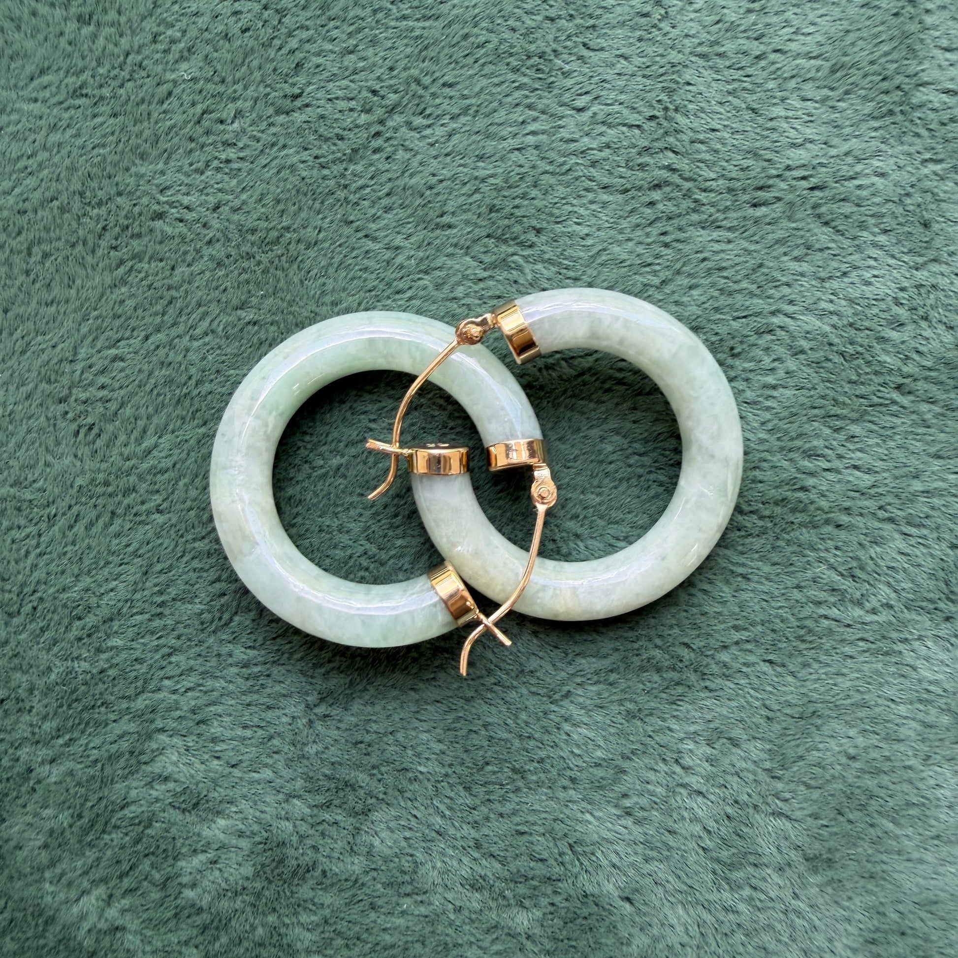 Two light green jade hoops with 14k gold hooks and end caps, displayed on a textured dark green surface.