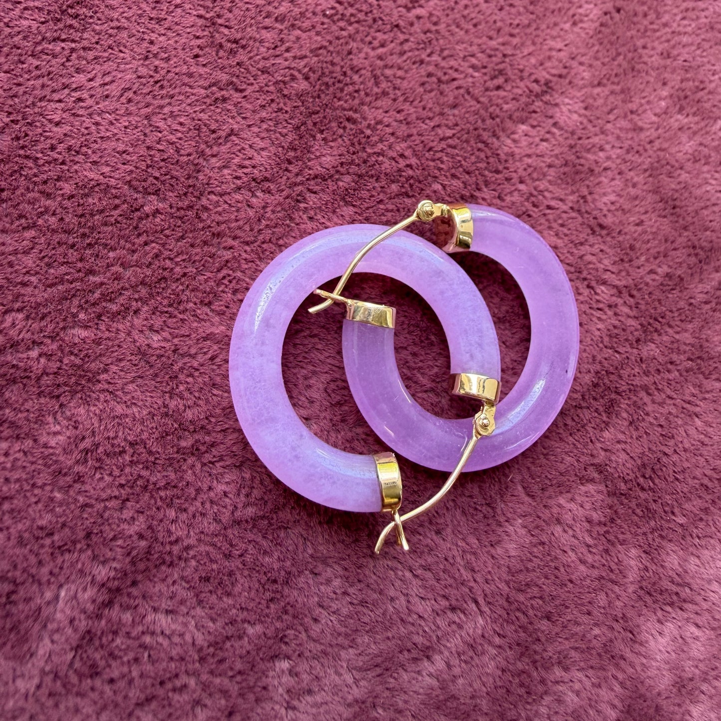 A pair of lavender jade hoop earrings with 14k gold hooks, displayed on a textured mauve fabric background.