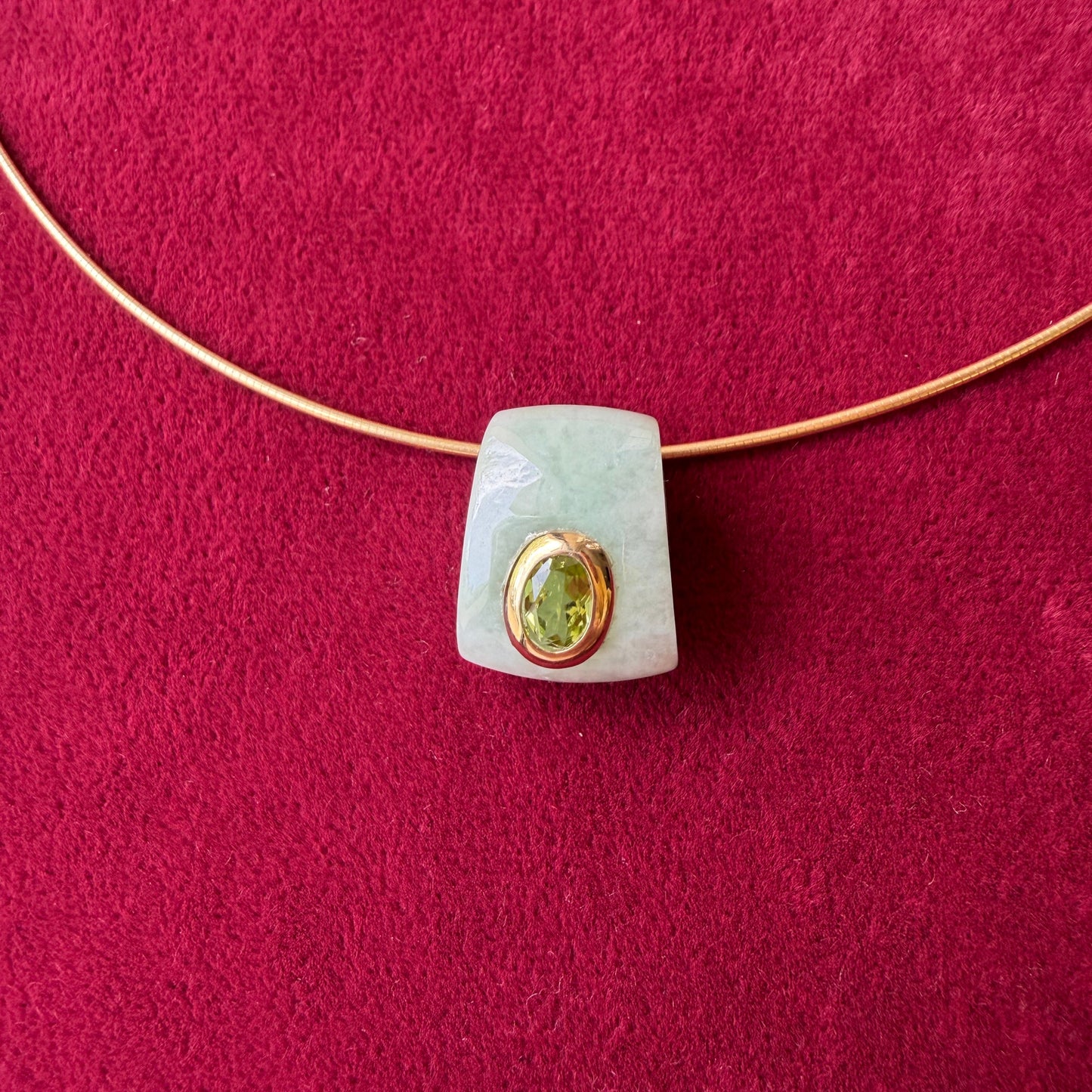 A light green jade pendant with a gold bezel-set oval peridot gemstone, threaded onto a gold necklace, displayed on a textured dark red background.