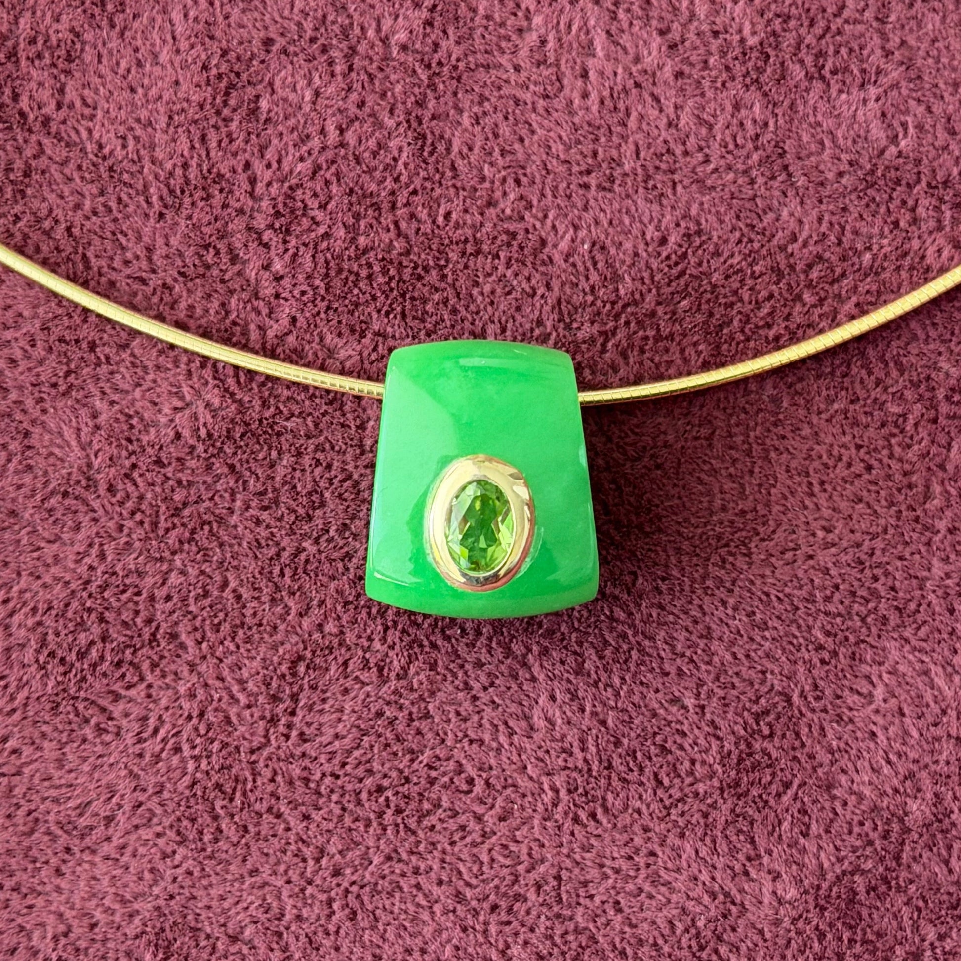 A close up view of a green jade and peridot pendant with a 14k gold bezel at the center, lying on a textured purple fabric background.