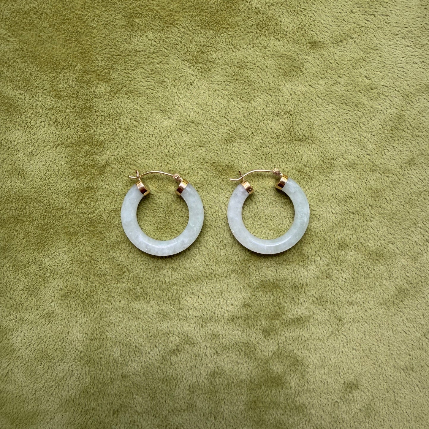 A pair of medium light green jade hoop earrings with 14k gold posts, displayed on a textured olive green surface.