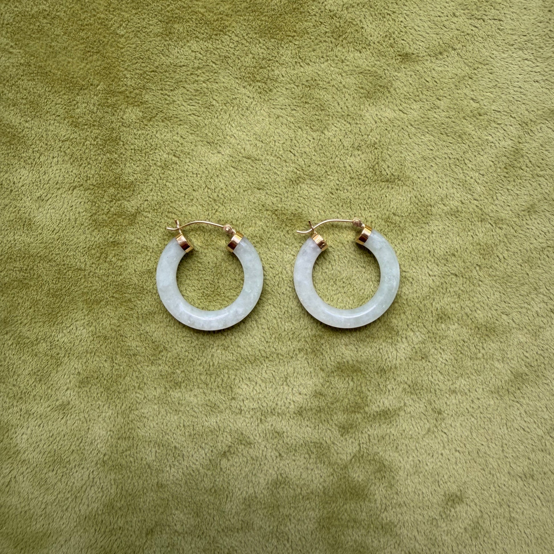 A pair of medium light green jade hoop earrings with 14k gold posts, displayed on a textured olive green surface.