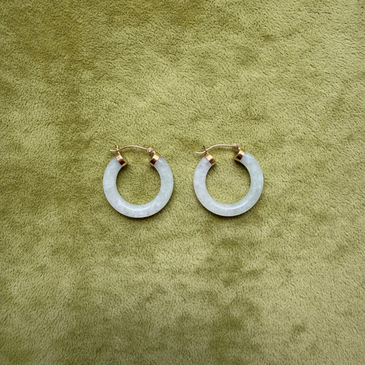 A pair of medium light green jade hoop earrings with 14k gold posts, displayed on a textured olive green surface.