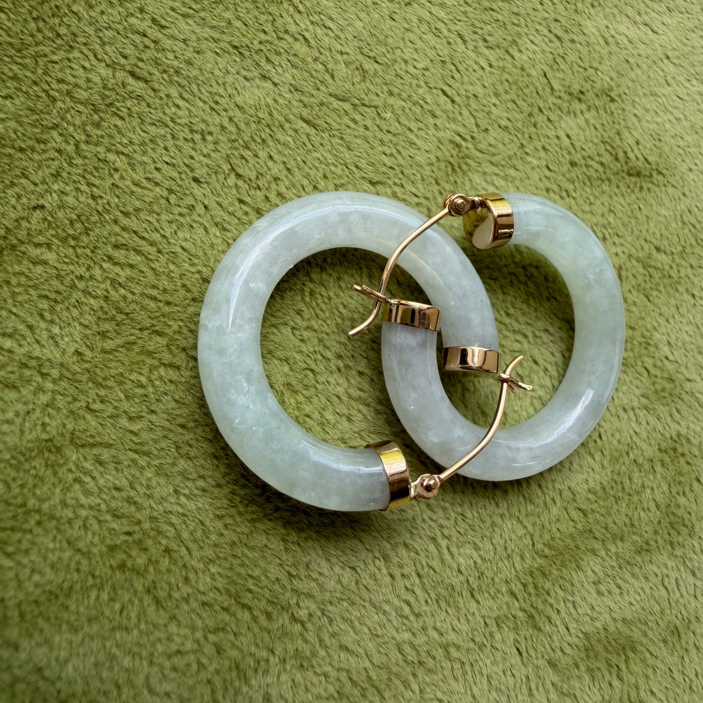 A pair of light green jade hoops with 14k gold hooks, displayed on a textured olive green fabric background.
