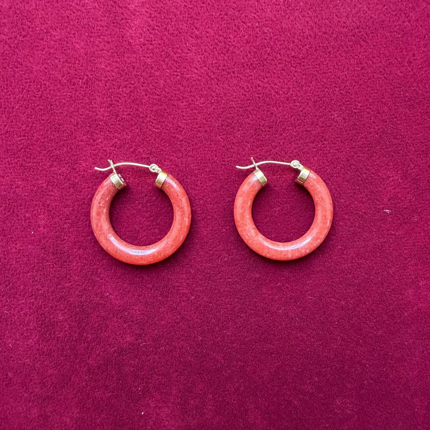 A pair of circular red jade hoop earrings with 14k gold metal clasps, displayed on a textured magenta background.