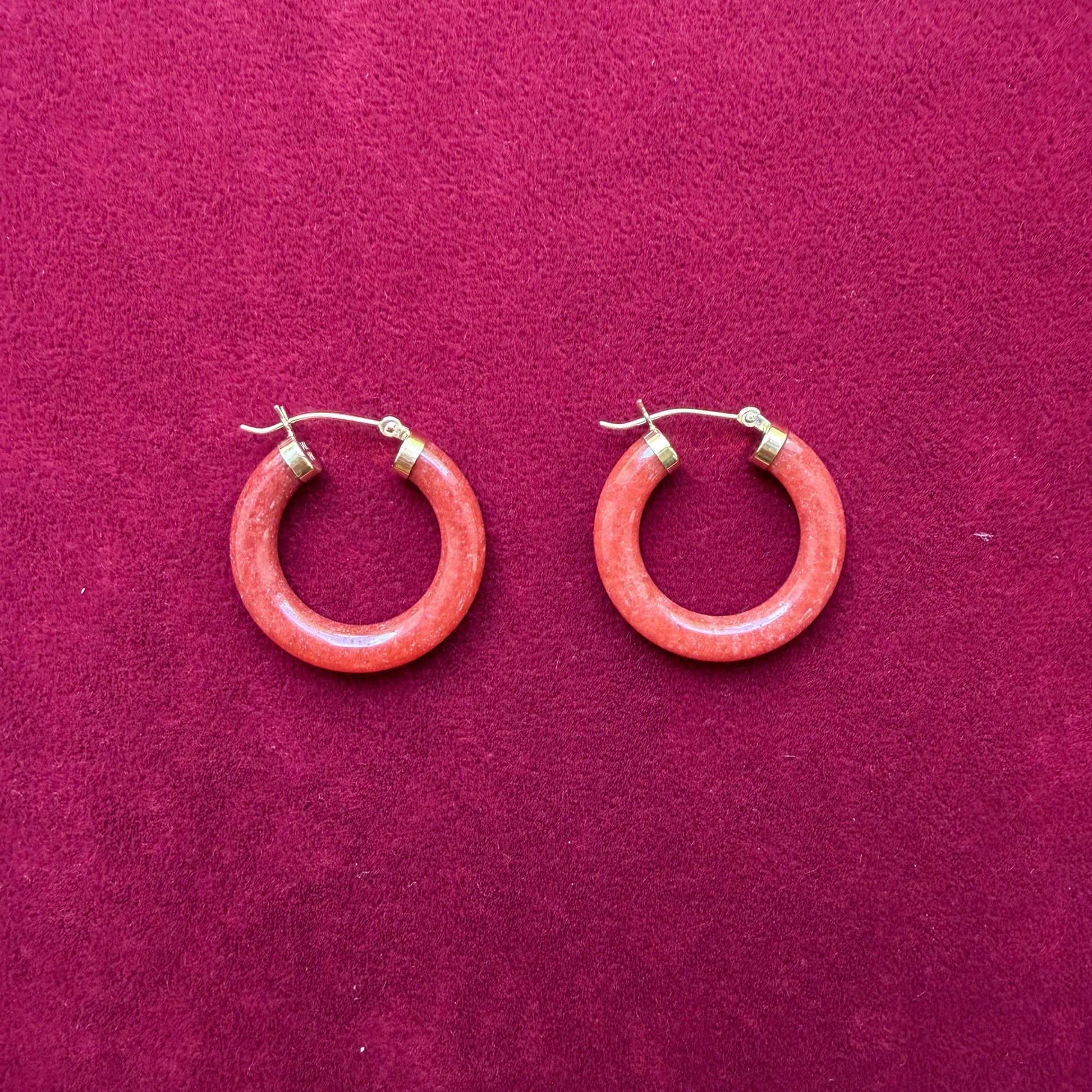 A pair of circular red jade hoop earrings with 14k gold metal clasps, displayed on a textured magenta background.
