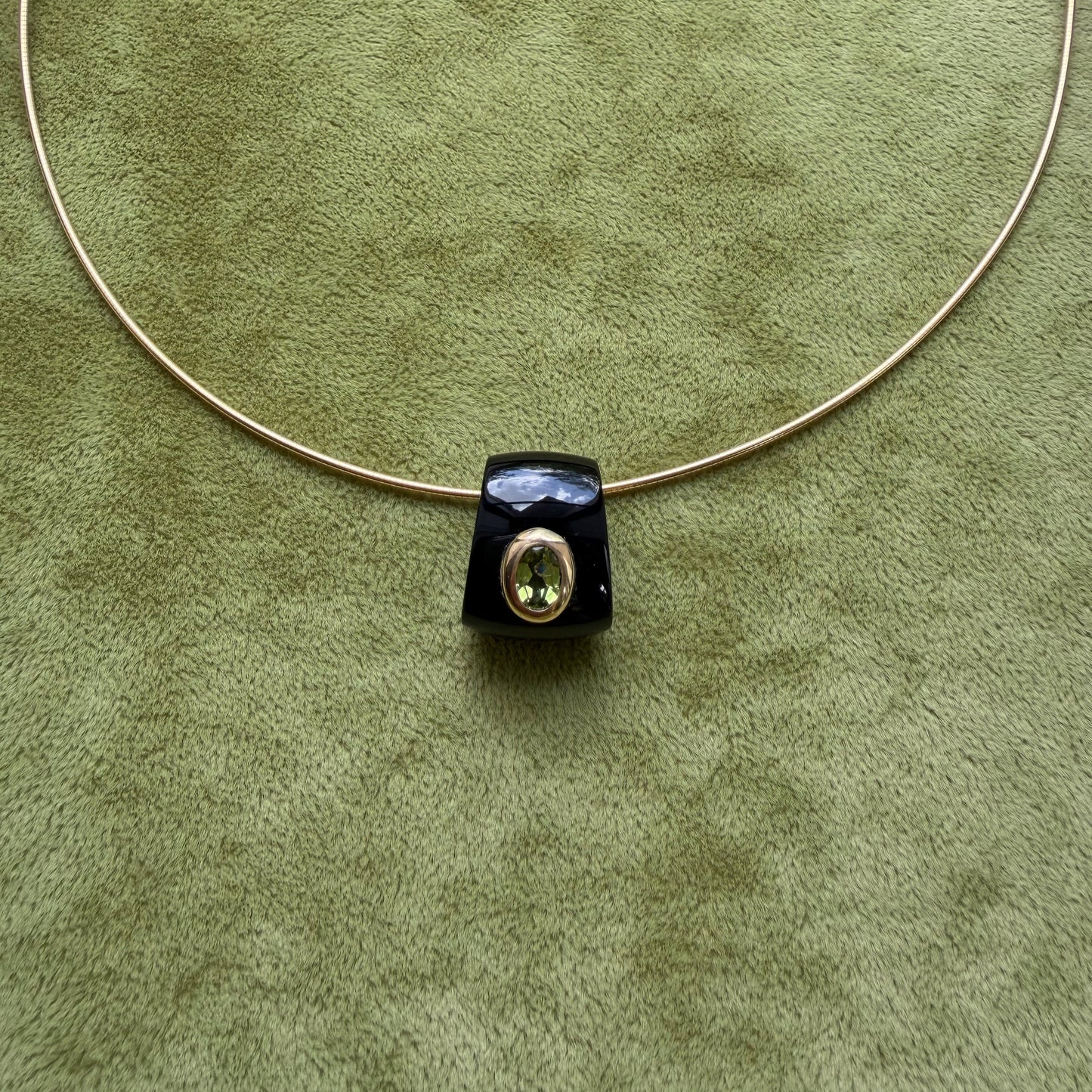 A black onyx pendant featuring a central peridot, displayed on a textured olive green background.