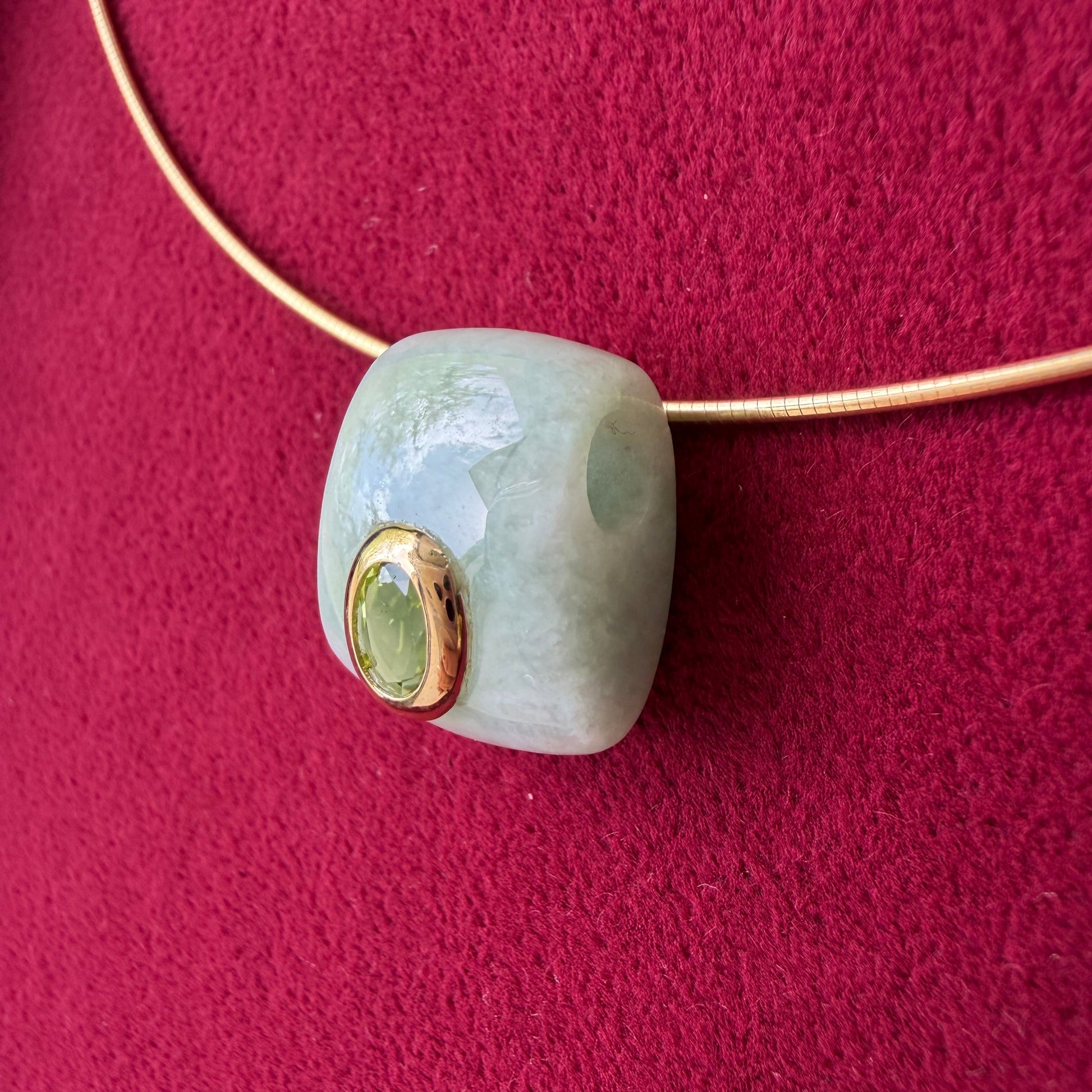 A light green jade cylindrical bead with a 14k gold bezel set peridot, shown on a gold necklace against a textured dark red background.
