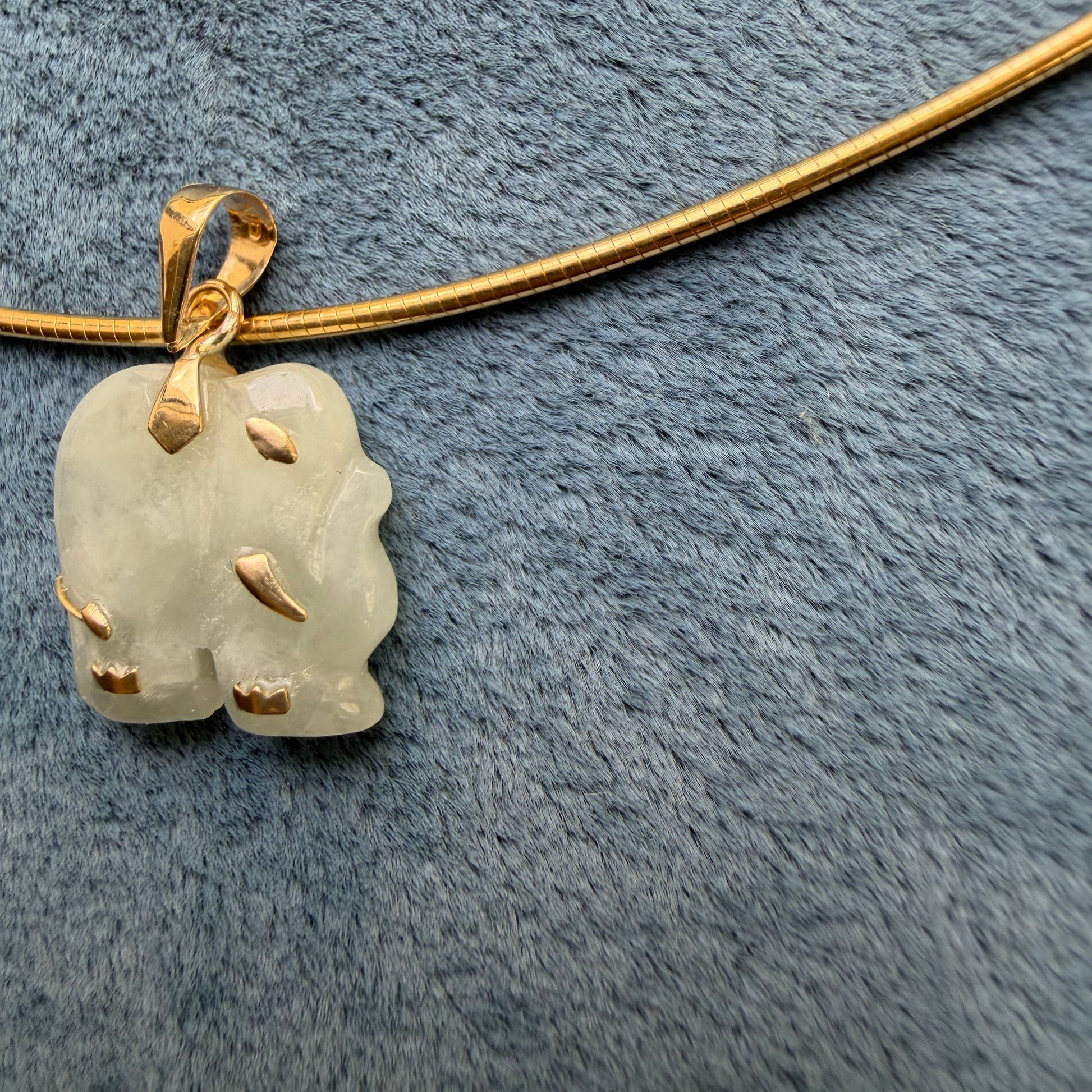 A 14k gold pendant necklace featuring an elephant carved, semi-translucent pale green jade charm, displayed on a textured blue-gray fabric background.