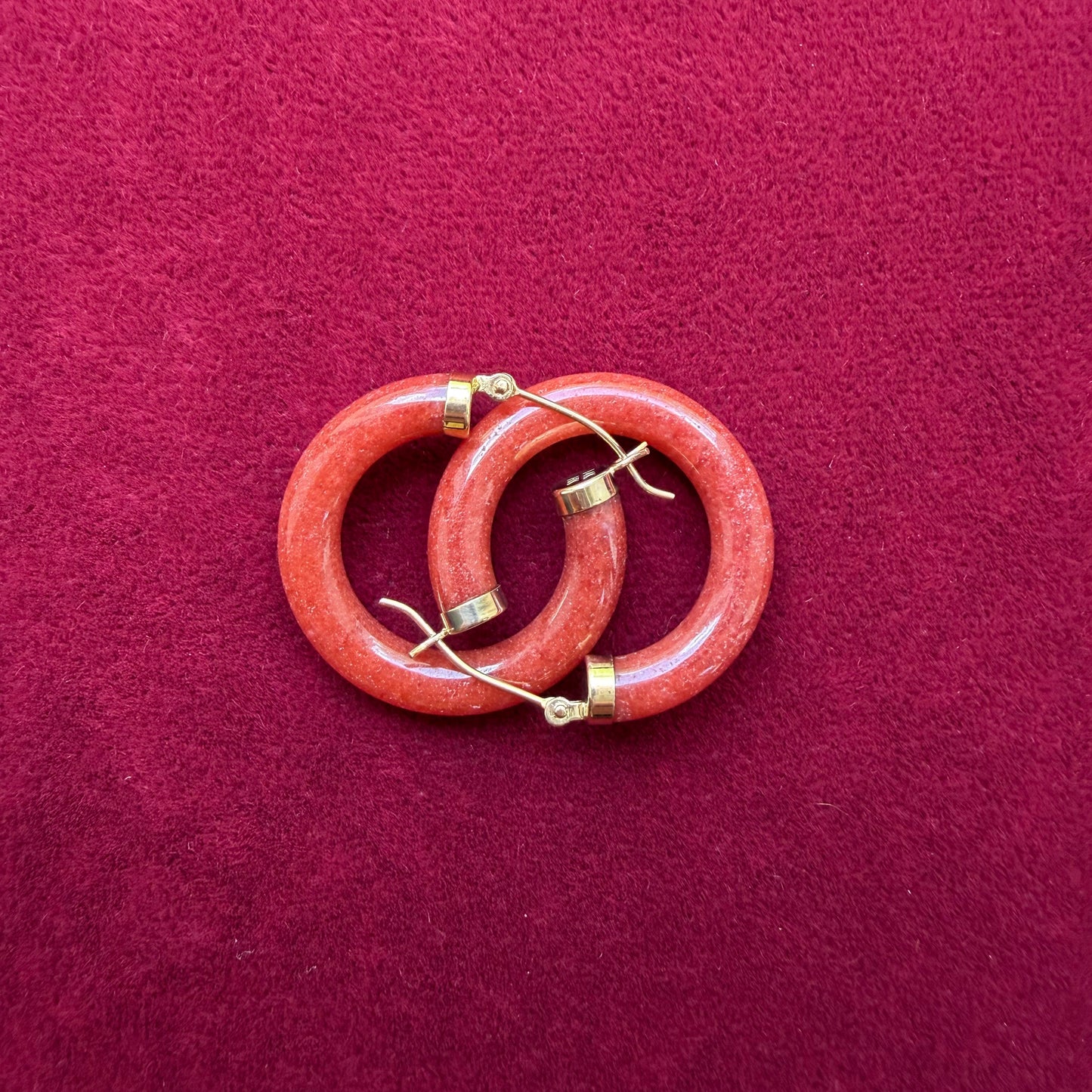 A pair of interlocking red jade hoops with 14k gold metal caps and hooks, displayed on a textured dark red background.