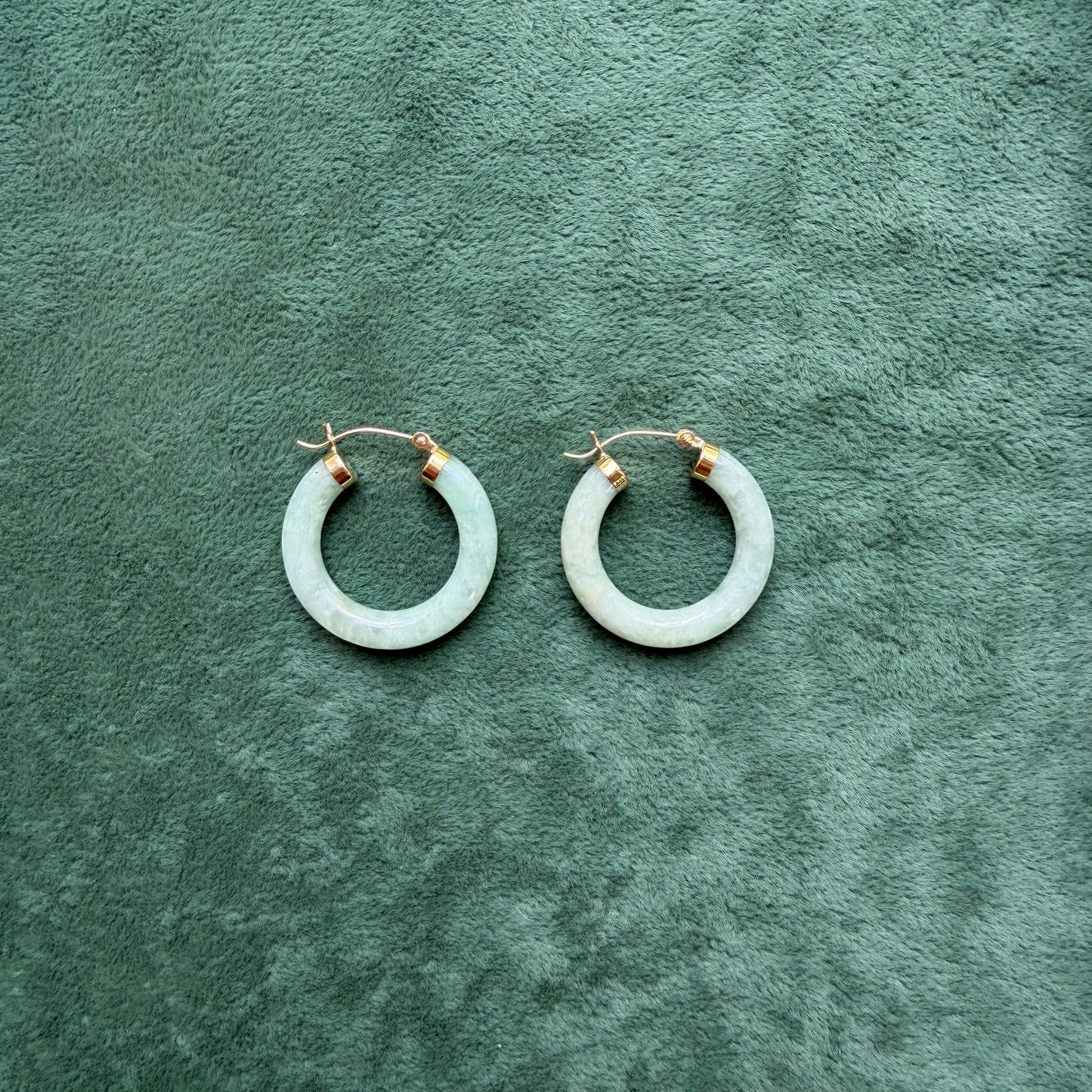 A pair of light green jade hoop earrings with 14k gold huggie clasps, displayed on a textured dark green velvet surface.