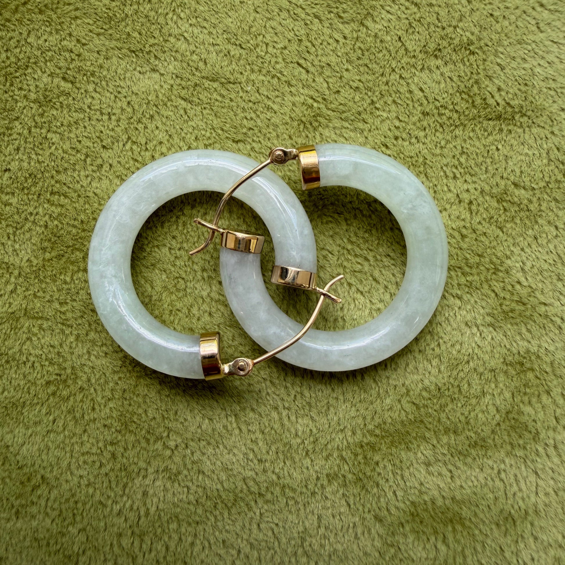 A pair of medium light green jade hoops with 14k gold ear loops and caps, displayed on a textured olive green fabric background.