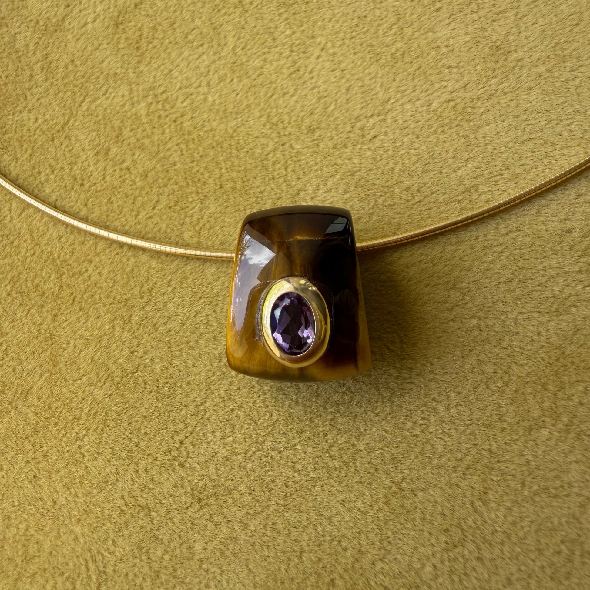 A tiger's eye pendant, featuring a bezel-set round purple amethyst center, resting on a textured olive green surface.