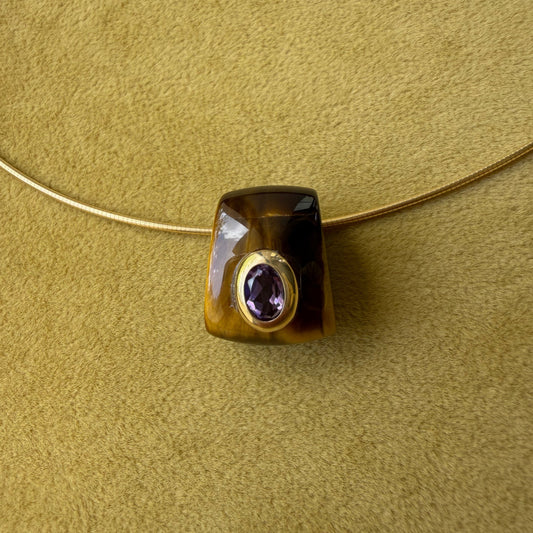 A tiger's eye pendant, featuring a bezel-set round purple amethyst center, resting on a textured olive green surface.