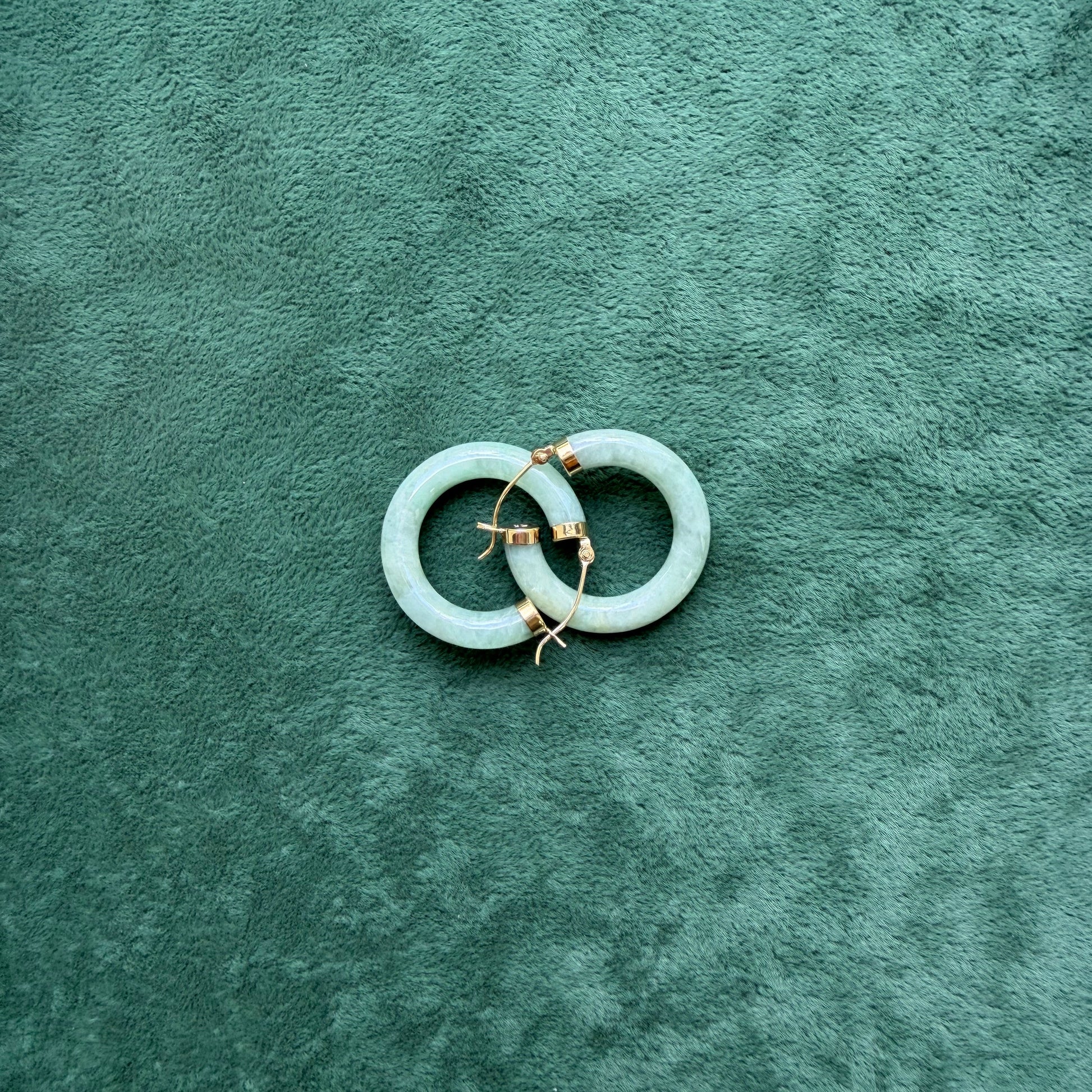 Two green jade hoops with 14k gold hooks and end caps, displayed on a textured dark green velvet surface.