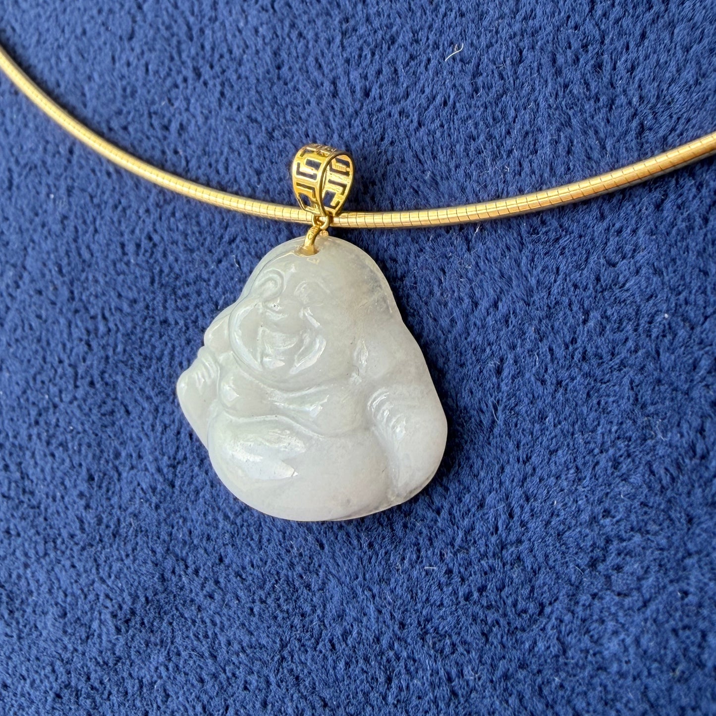 A carved white jade buddha pendant with a 18k gold bail, displayed on a textured blue background.