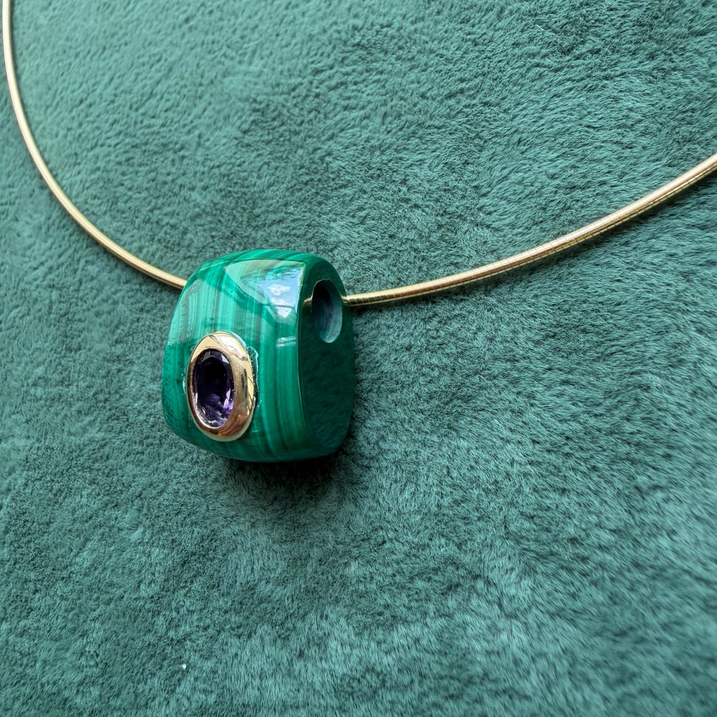 A green malachite pendant with a 14k bezel-set purple amethyst center, threaded onto a gold necklace against a textured green background.