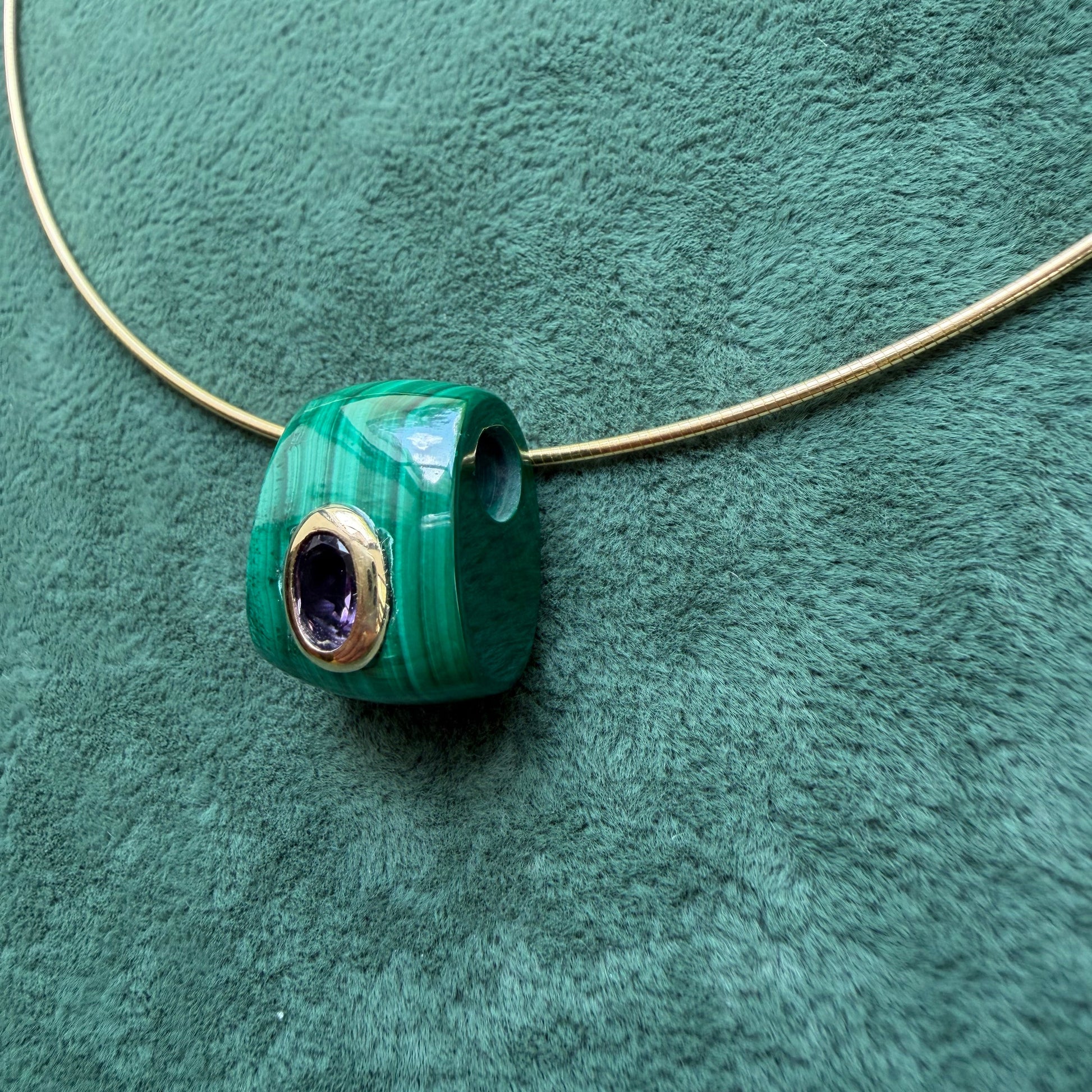 A green malachite pendant with a 14k bezel-set purple amethyst center, threaded onto a gold necklace against a textured green background.