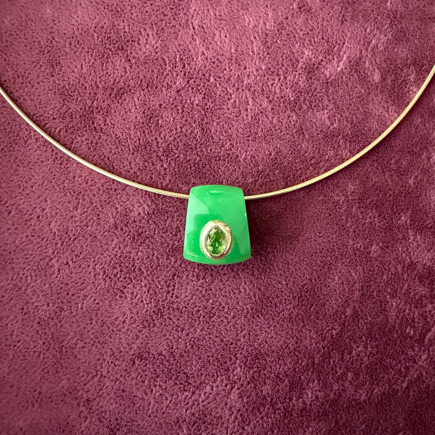 A green jade pendant with a 14k solid gold bezel hugging a peridot stone, threaded onto a thin gold wire, set against a textured dark purple background