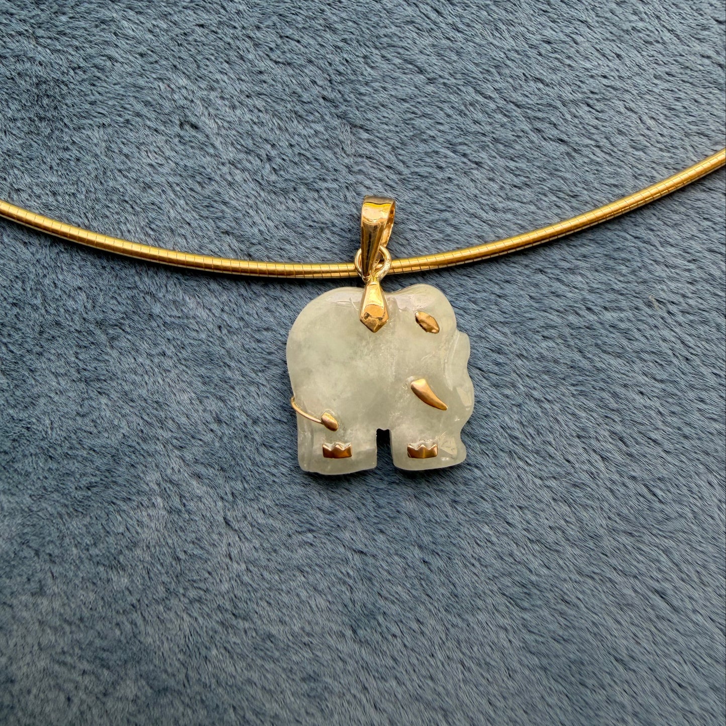 A small, light green jade carved elephant pendant, displayed on a textured blue-gray fabric background.