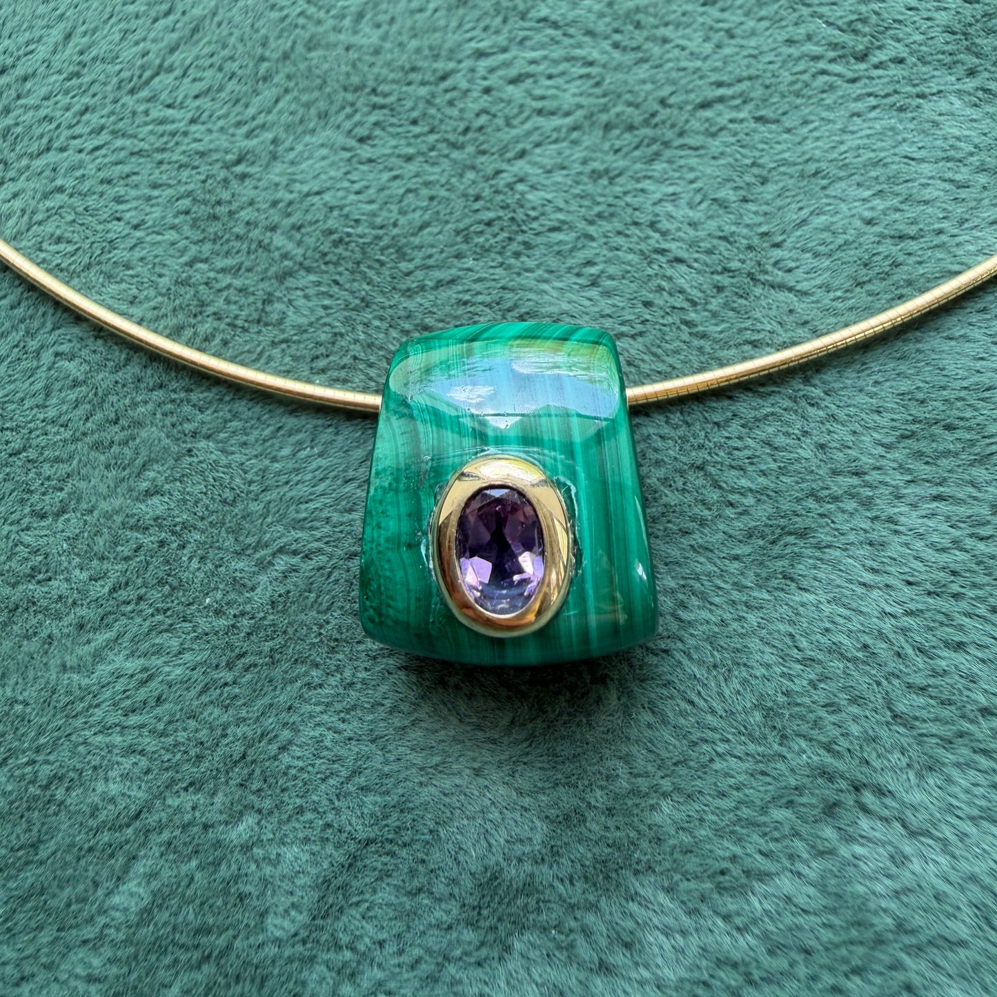 A malachite pendant with and a central oval amethyst gemstone, displayed on a textured green background.