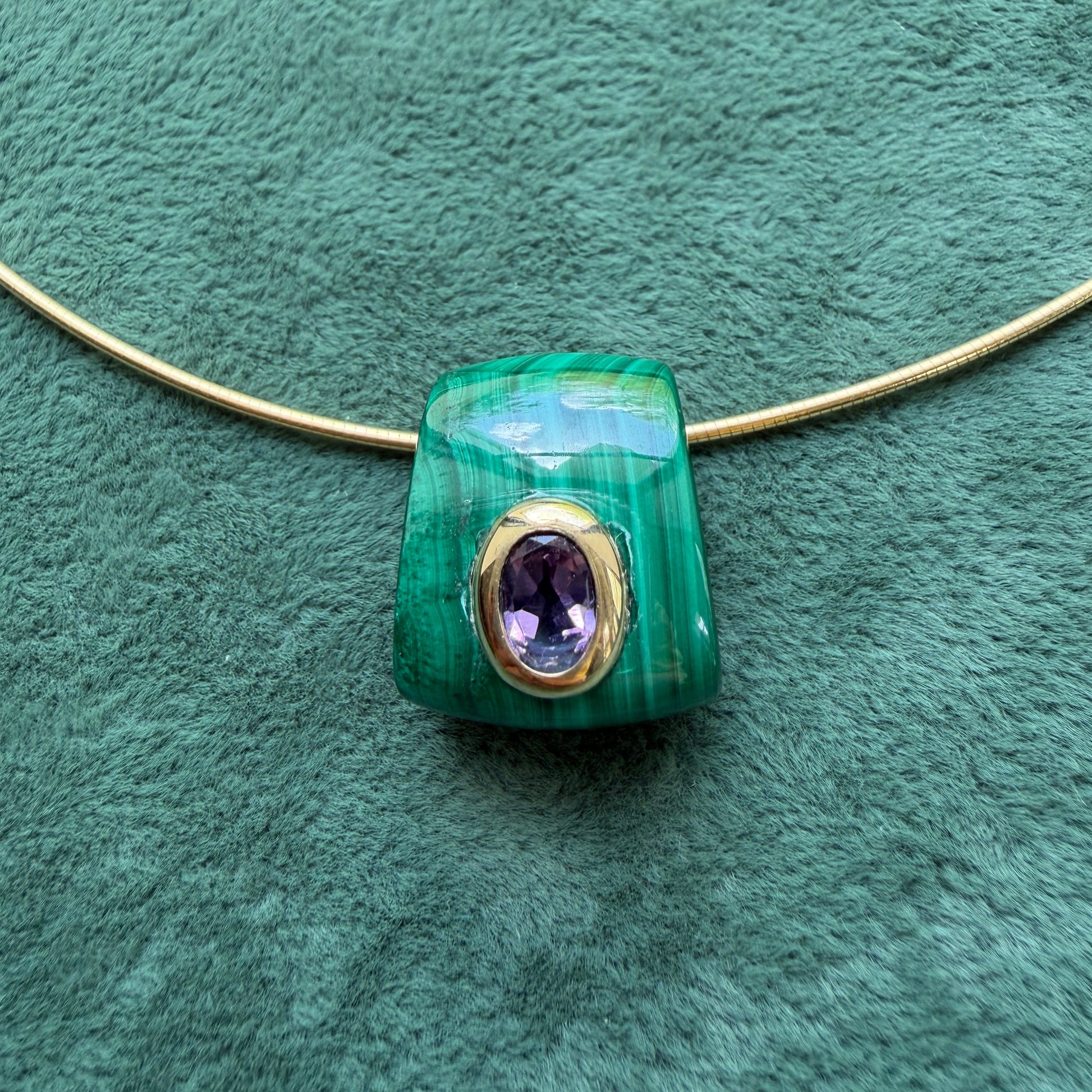 A malachite pendant with and a central oval amethyst gemstone, displayed on a textured green background.