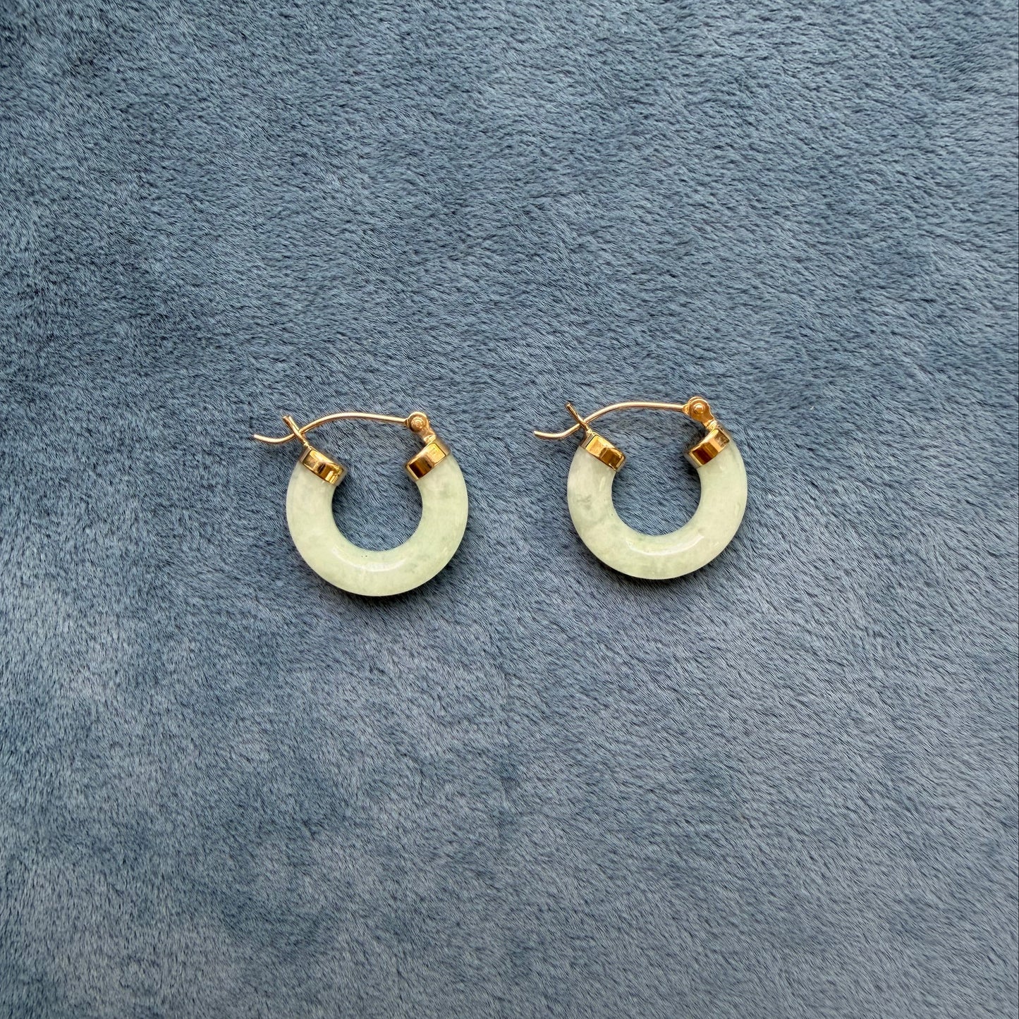 A pair of small 14k gold light green jade hoop earrings, displayed on a textured gray fabric background.