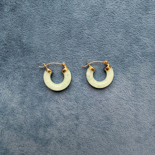 A pair of small 14k gold light green jade hoop earrings, displayed on a textured gray fabric background.
