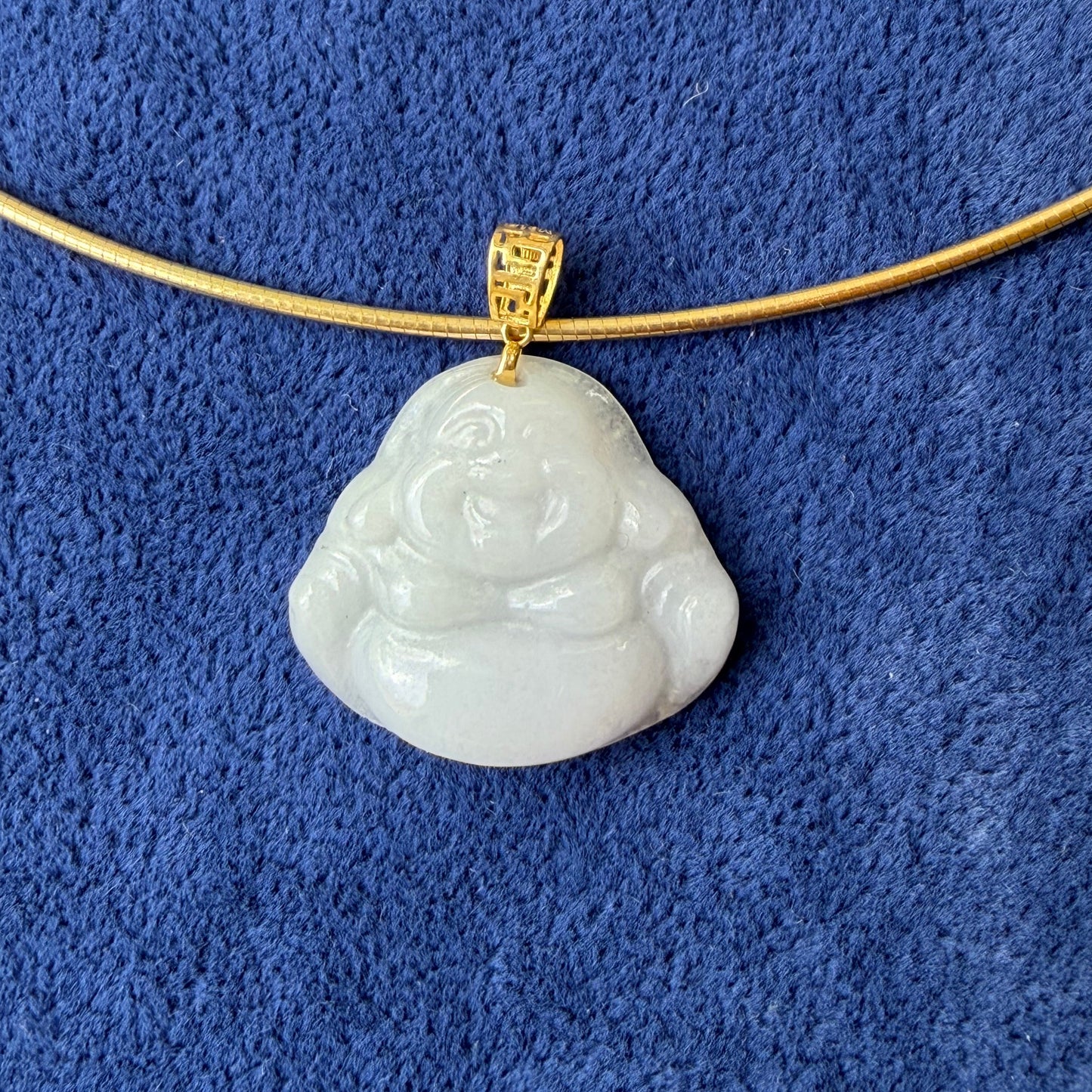 A carved white jade Buddha pendant on a gold chain, displayed on a textured blue fabric background.