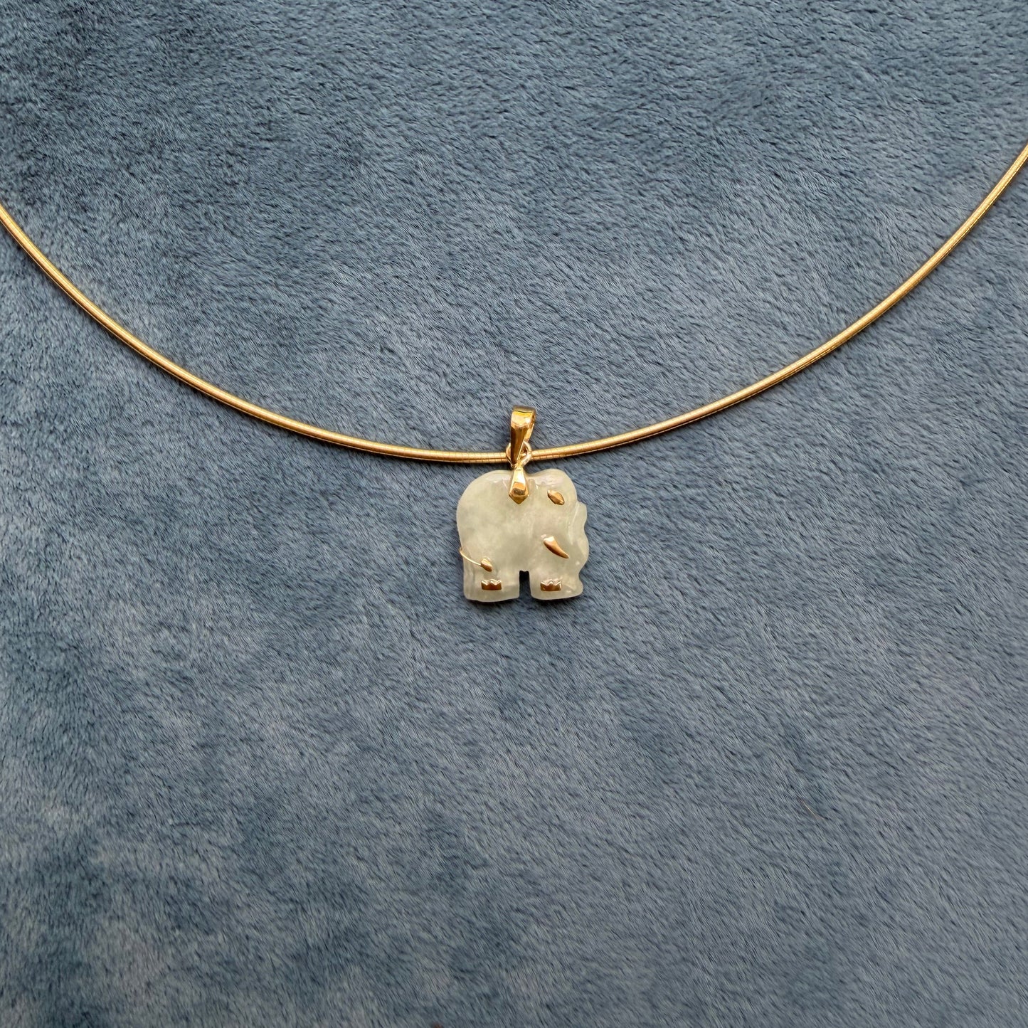 A delicate gold wire necklace with a small, light green jade carved elephant pendant, laid flat on a textured blue-gray fabric background.