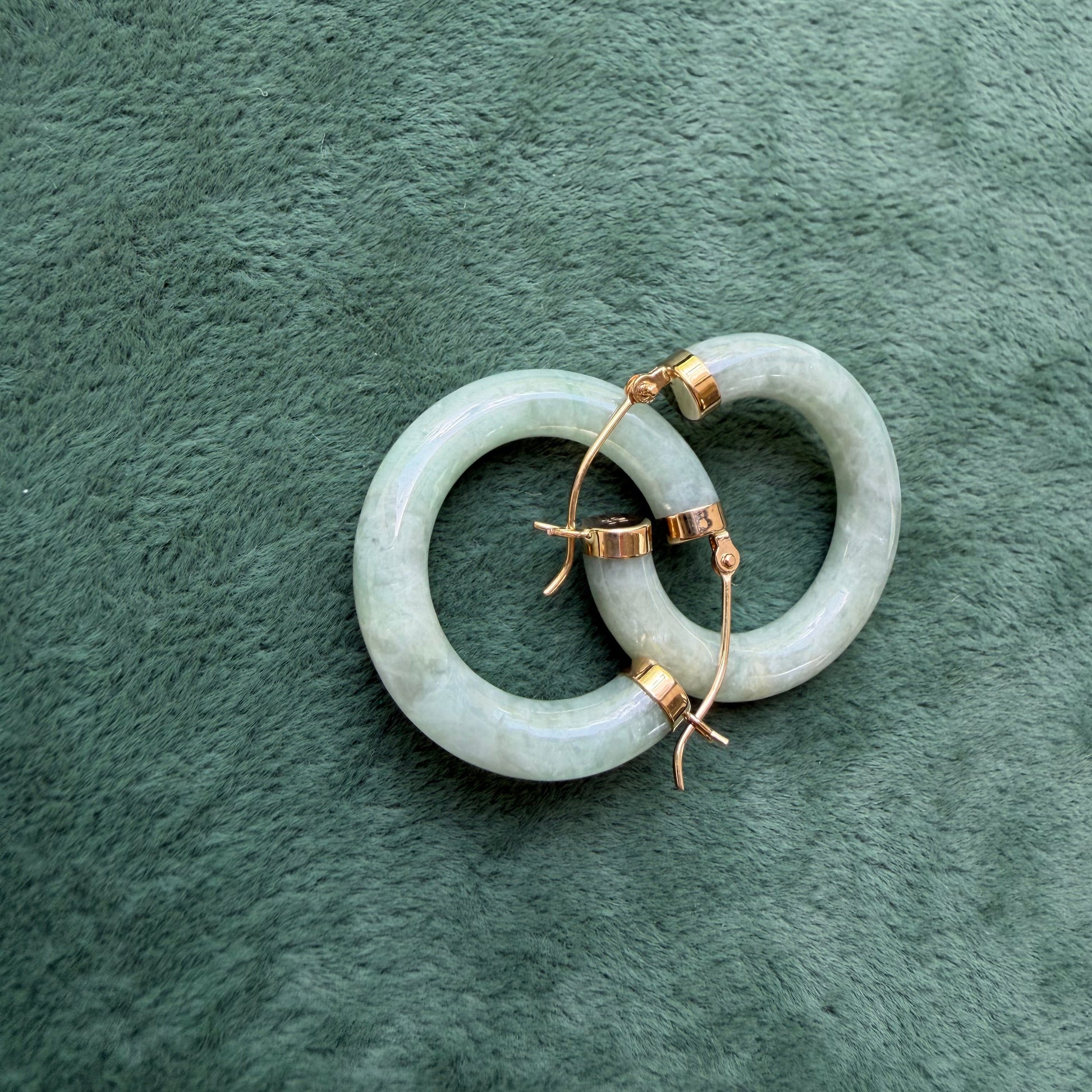 Two green jade hoops with 14k gold hooks and end caps, displayed on a textured dark green fabric background.