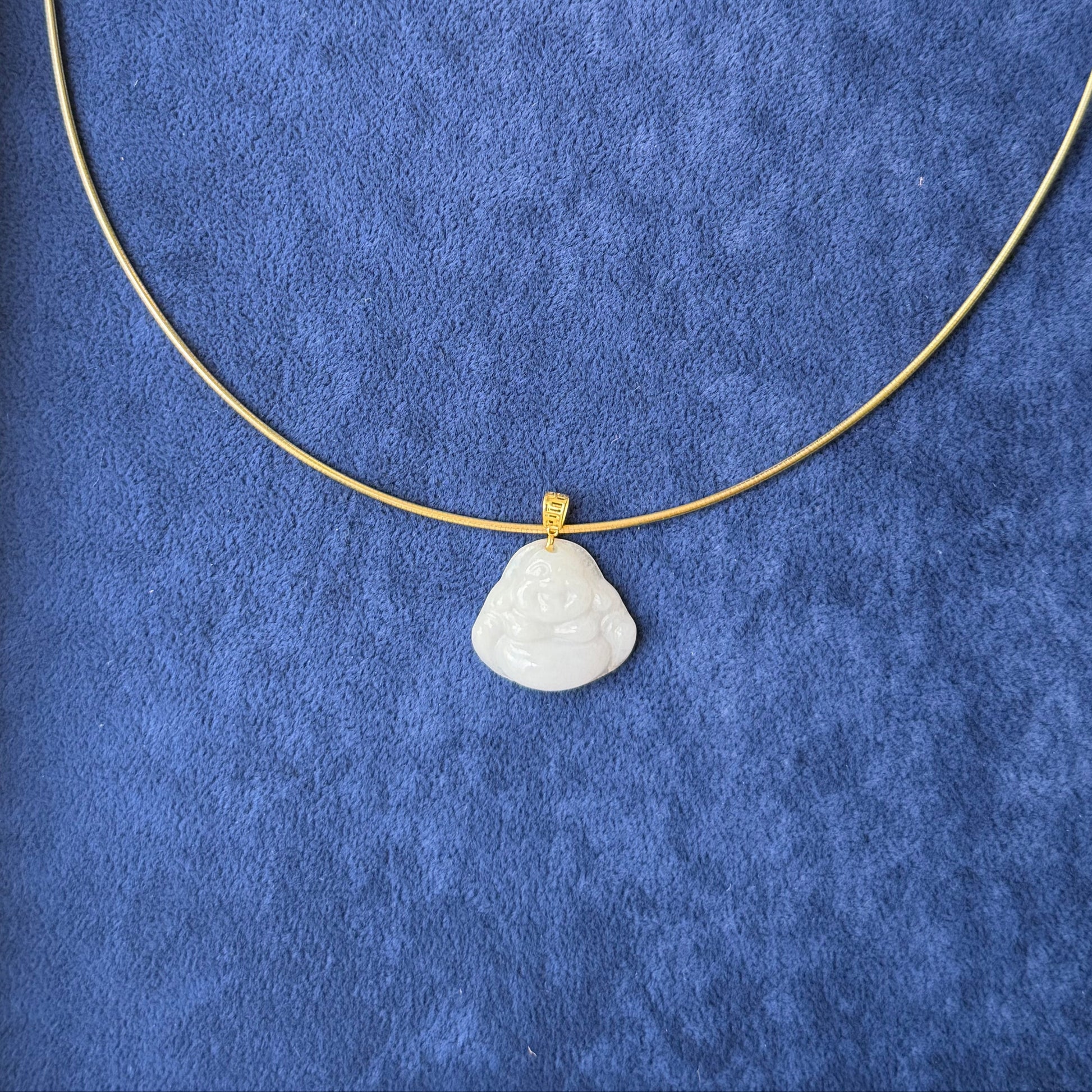 A delicate gold necklace with a small, white jade buddha pendant, displayed on a textured blue velvet jewelry box insert.
